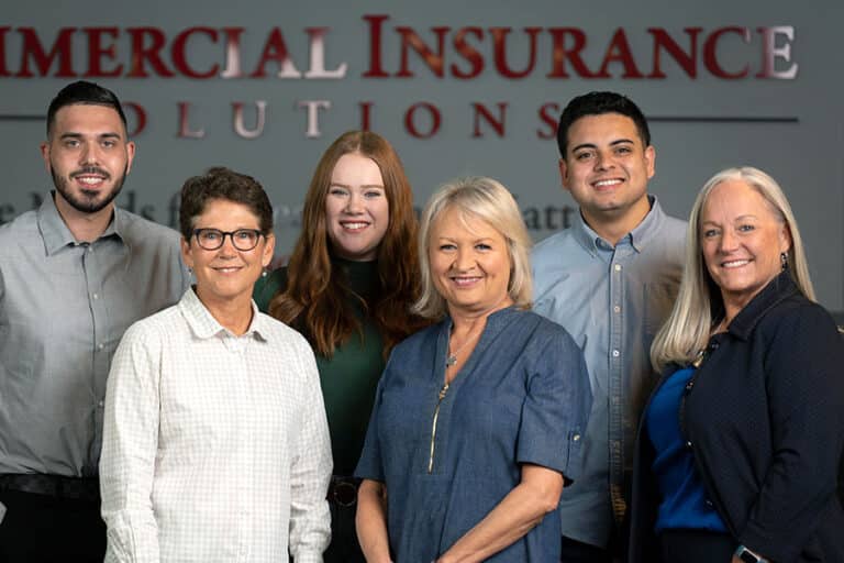 Group of six smiling people, three men and three women, standing in front of a wall with the text "Commercial Insurance Solutions.