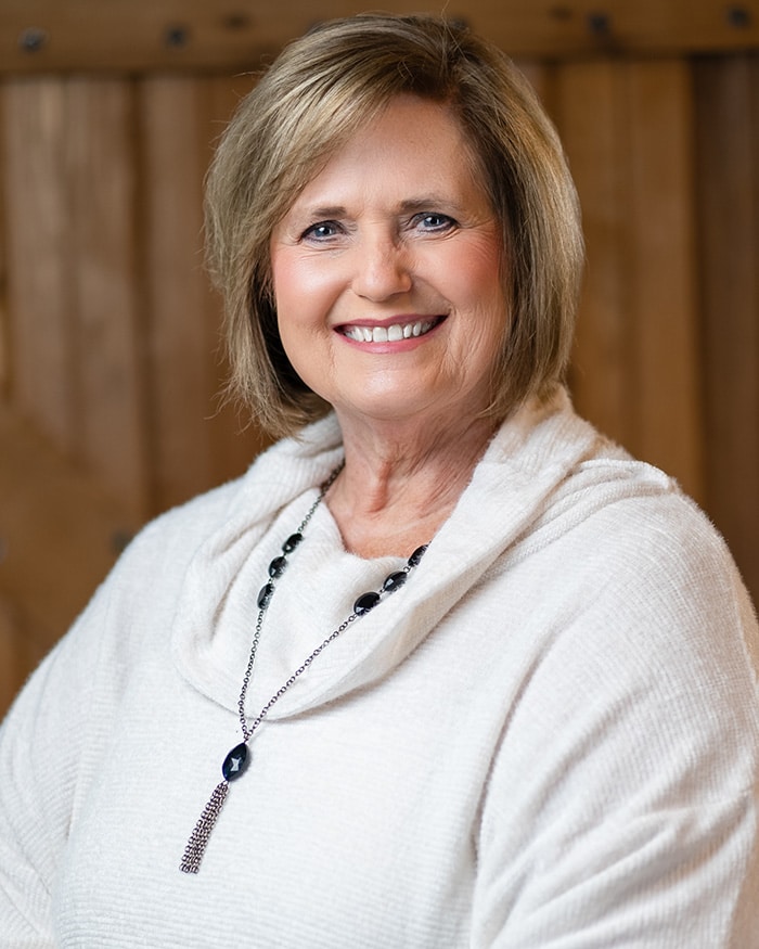 A woman with short blonde hair smiles while wearing a light-colored sweater and a long necklace, standing in front of a wooden backdrop.