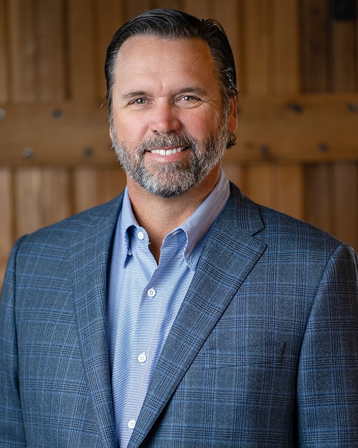 Man with graying beard and hair wearing a blue button-up shirt and checkered blazer, smiling at the camera with a wooden wall background.