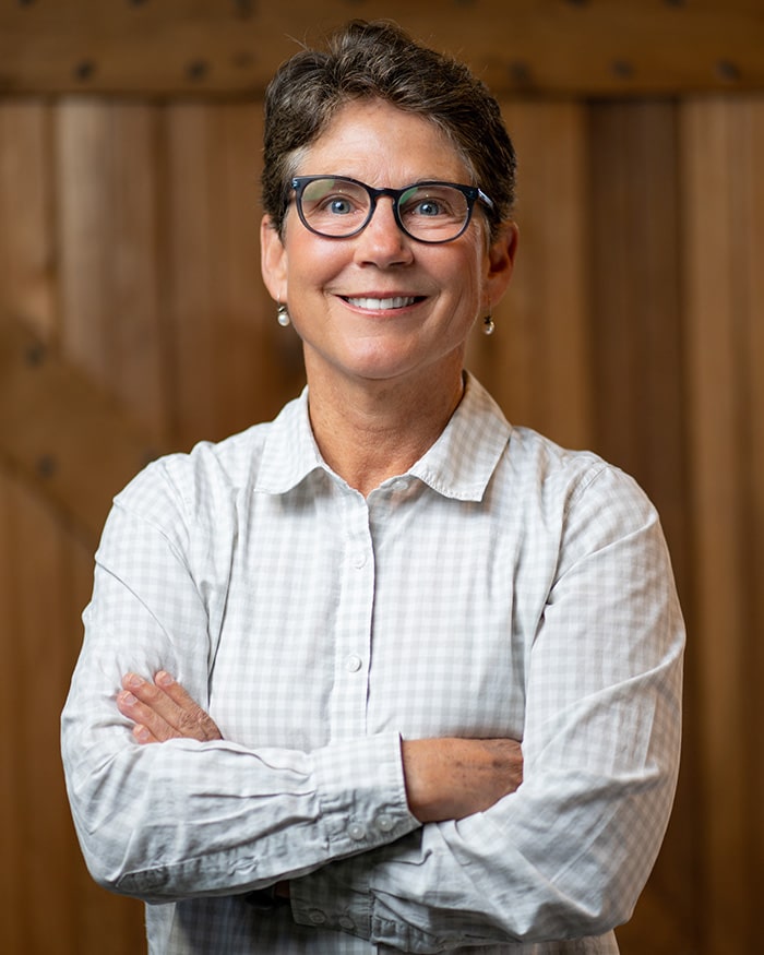 Person with short hair, glasses, and a white checkered shirt standing with arms crossed in front of a wooden background.