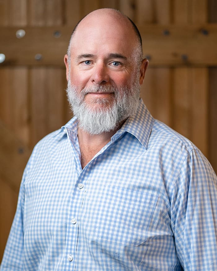 A man with a white beard is wearing a blue and white checkered shirt, standing in front of a wooden background.