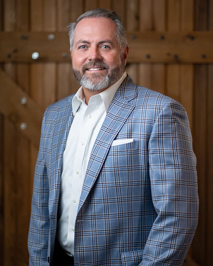 A man with short gray hair and a beard, wearing a light blue plaid suit jacket over a white shirt, stands smiling in front of a wooden background.