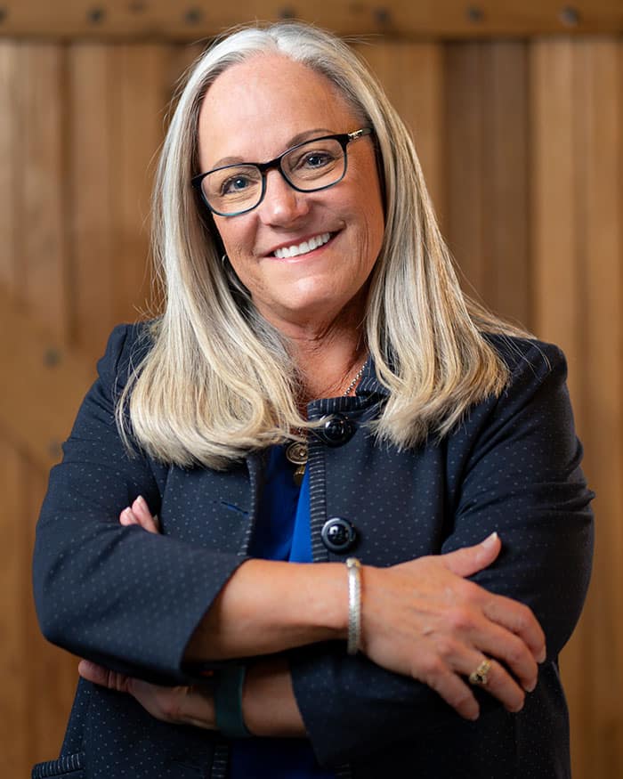 A woman with long gray hair and glasses smiles while crossing her arms. She is wearing a dark blazer and standing in front of a wooden background.