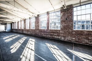 An empty industrial-style room with large windows, exposed brick walls, and natural light streaming through.