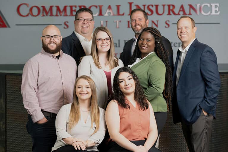 A group of eight professionally dressed individuals poses together in front of a sign that reads “Commercial Insurance Solutions.”.