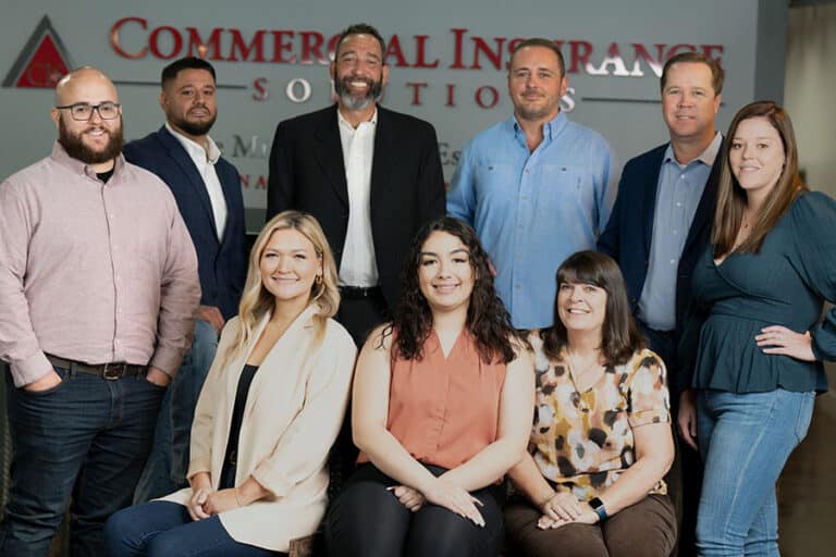 A group of eight people posing for a photo in front of a Commercial Insurance sign. Five are standing, and three are seated. They are all dressed in business casual attire.