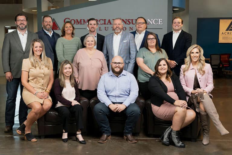 A group of 14 people, dressed in business casual attire, pose for a group photo in an office setting with a sign reading "Commercial Insurance Solutions" in the background.