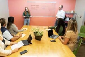 A woman stands at a whiteboard giving a presentation to a group of five people seated at a table in a conference room.