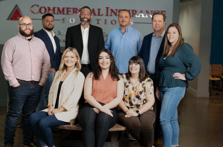 A group of nine people posed for a group photo in front of a "Commercial Insurance" sign. Four are seated in front while five stand in the back. The setting is an office environment.