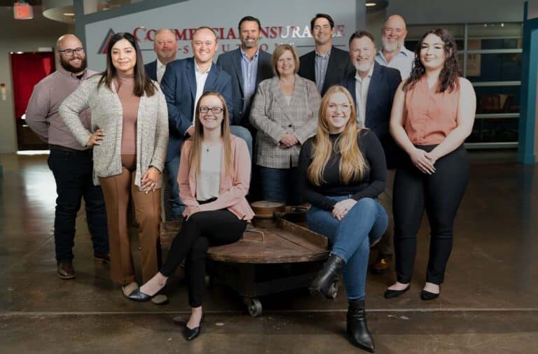 A group of eleven people, casually dressed, stand and sit together inside an office building, posing for a photo in front of a wall with partially visible text.