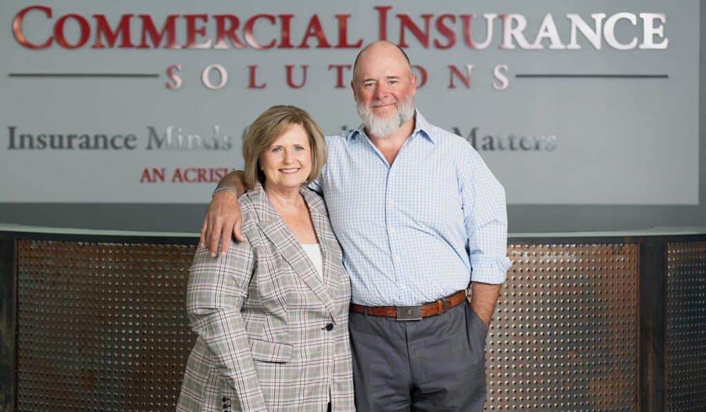 Two individuals, one woman and one man, standing together in front of a sign reading "Commercial Insurance Solutions" in an office setting. The man has his arm around the woman.