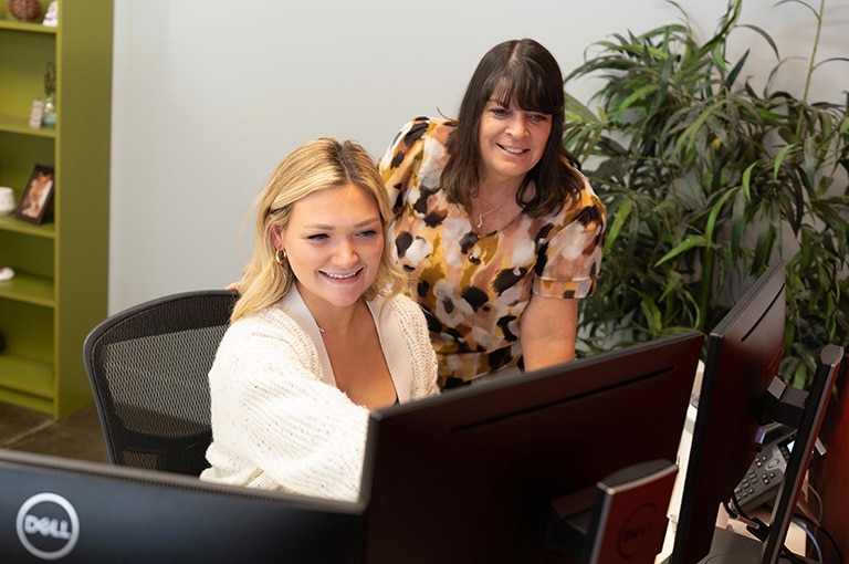 Two women are at a desk looking at a computer monitor. One is sitting and using the computer, while the other stands behind her, both smiling. There is a potted plant and a bookshelf in the background.