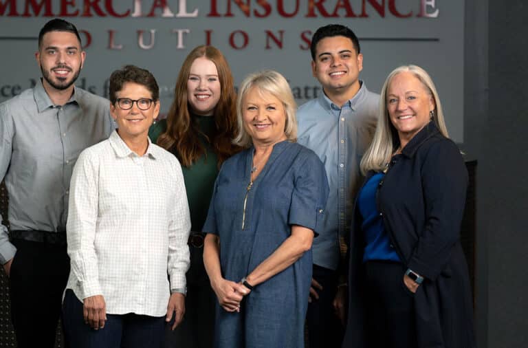 A group of six people standing and smiling in front of a sign that reads 'Commercial Insurance Solutions.'.