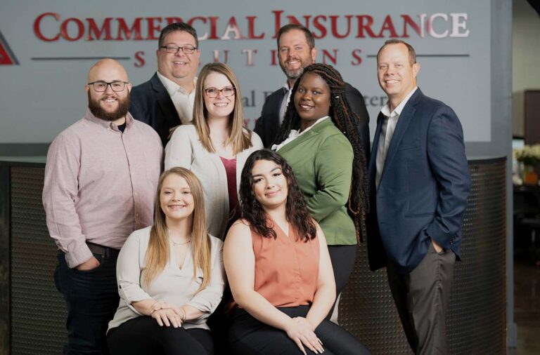 A group of eight people, five standing and three sitting, posing together for a photo in an office setting. The backdrop includes a sign that reads "Commercial Insurance Solutions".