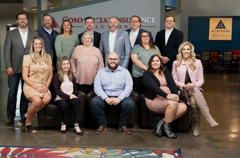 A group of 14 people are posing for a group photo in an office setting. Some are standing while others are seated on chairs, with a company logo “Commercial Insurance Solutions” visible in the background.