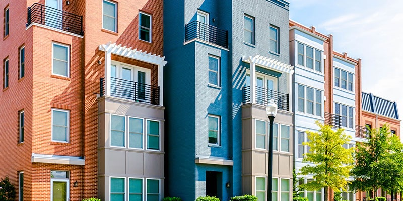 A row of modern, colorful townhouses with multiple floors, balconies, and varied facades in a residential area, with a streetlamp and trees in front.