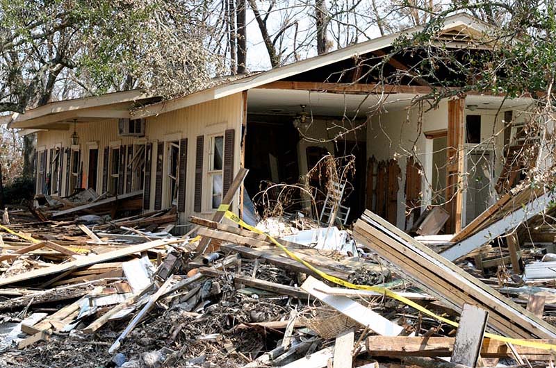 A single-story house with significant structural damage, partially collapsed walls and roof, surrounded by debris and fallen tree branches.