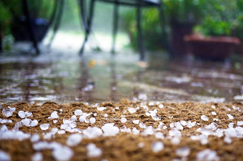 Hailstones scattered on a doormat, with a blurred background showing outdoor furniture and a garden, during a hailstorm.