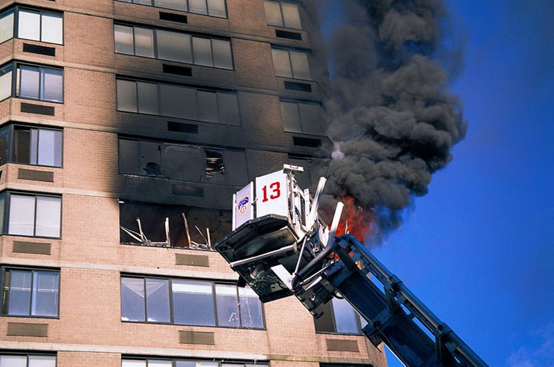 A fire truck ladder extends to a high-rise building with visible flames and heavy black smoke emanating from broken windows.